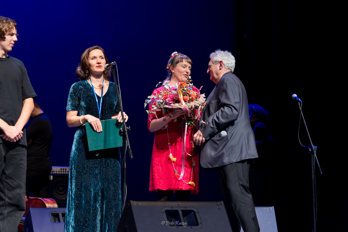 Anna Czapnik-Wójcik mówi do mikrofonu, obok Janusz Olszyński wręcza bukiet kwiatów Hance.