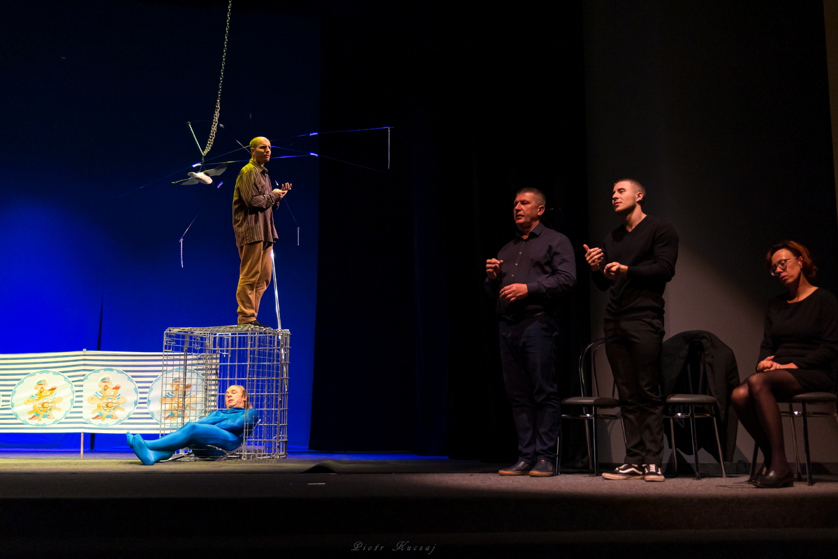 Na scenie mężczyźni – jeden leży na leżaku ustawionym wewnątrz metalowej klatki, drugi stoi na klatce ponad nim. Dwaj tłumacze migają, tłumaczka siedzi.