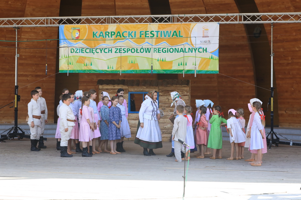 Fotografia: na scenie amfiteatru występuje grupa dzieci w ludowych strojach. Nad nimi baner z napisem: Karpacki Festiwal Dziecięcych Zespołów Regionalnych.