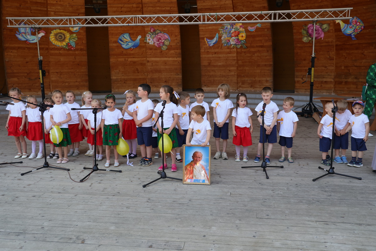 Dzieci ubrane w biało-czerwone i biało-niebieskie stroje. Przed nimi portret Jana Pawła II.