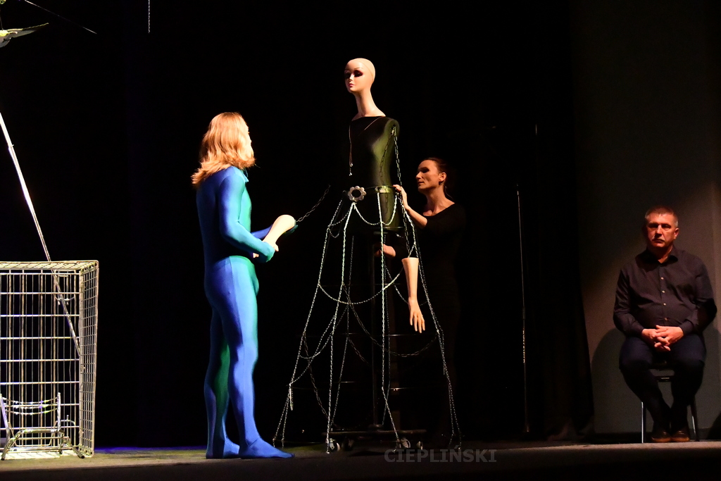 Scena ze spektaklu,mężczyzna w niebieskim kombinezonie, kobiecy manekin i kobieta, po prawej stronie siedzi tłumacz.