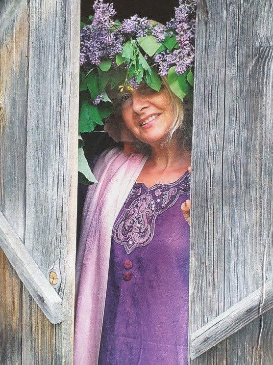 Małgorzata Ewa Czernik ubrana w jasnofioletową sukienkę z szalem. Na głowie ma duży wieniec z gałęzi bzu o liliowych kwiatach. Stoi między skrzydłami drewnianych drzwi.