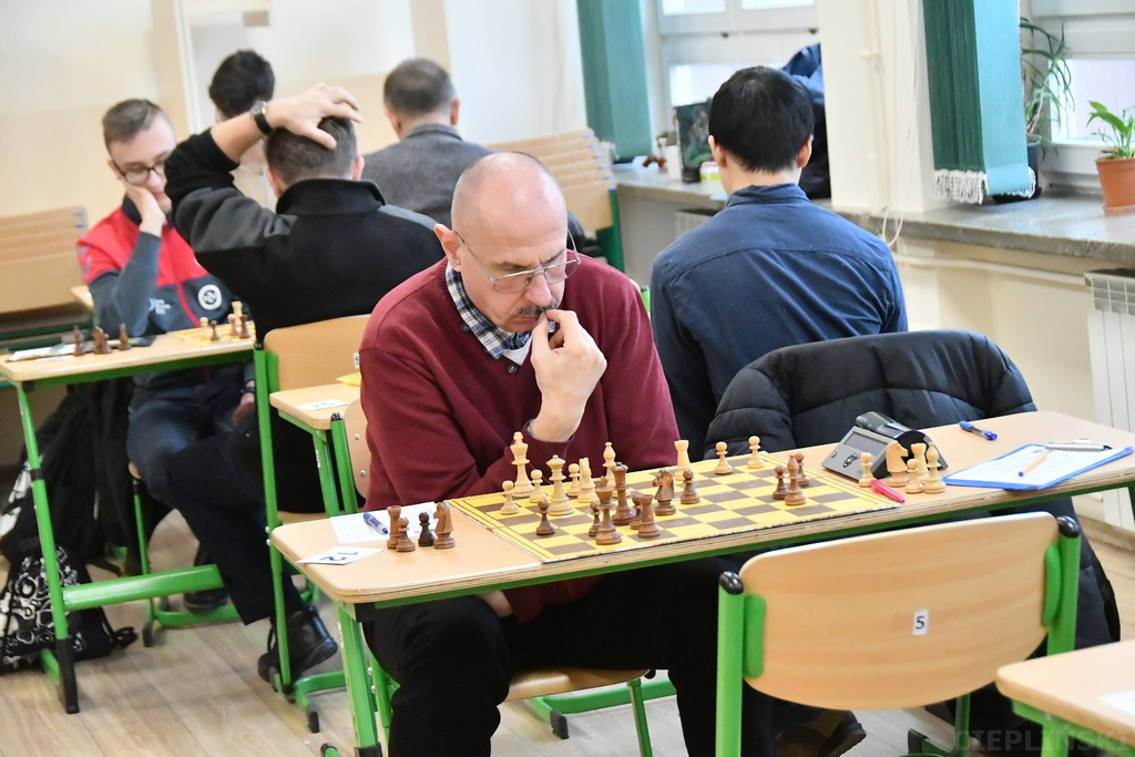 Mężczyzna samotnie pochyla się zamyślony nad szachownicą. W tle inni gracze.